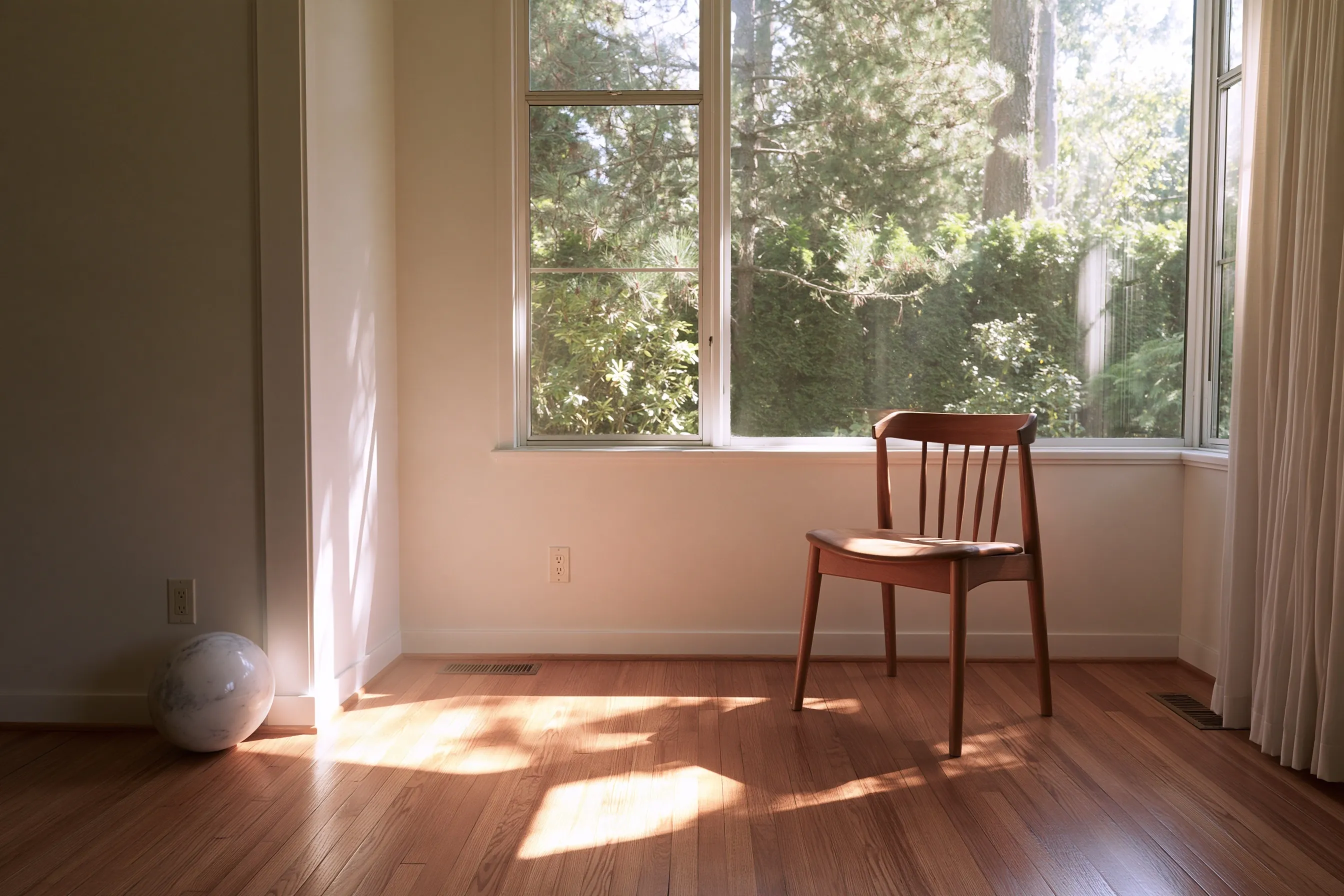 An empty living room with a hardwood floor, a wooden chair by a large window onto trees and a small ball resting against the wall in the corner